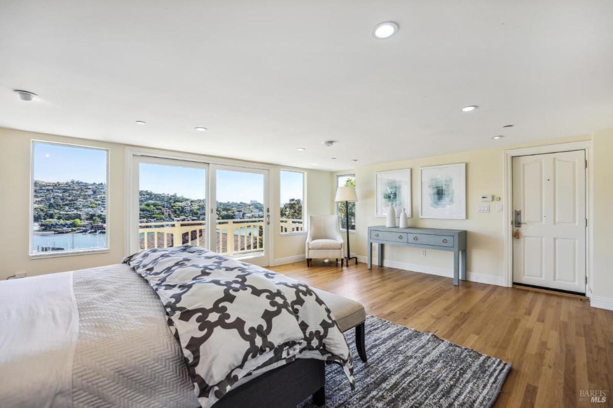 A bedroom with large windows overlooking the water, a patterned bedspread, and light wood flooring.