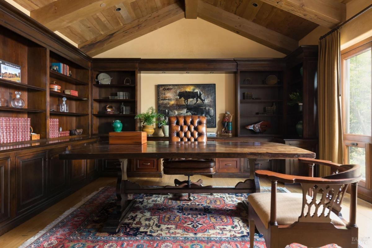 Office featuring dark wooden shelves filled with books and decor, a large wooden desk with a leather chair, and a patterned rug on the floor.