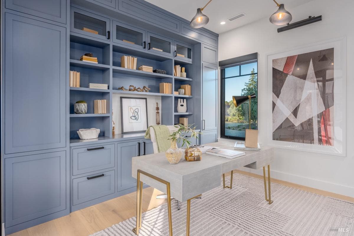 A home office with built-in blue cabinetry, a large desk with brass legs, and a window providing natural light.