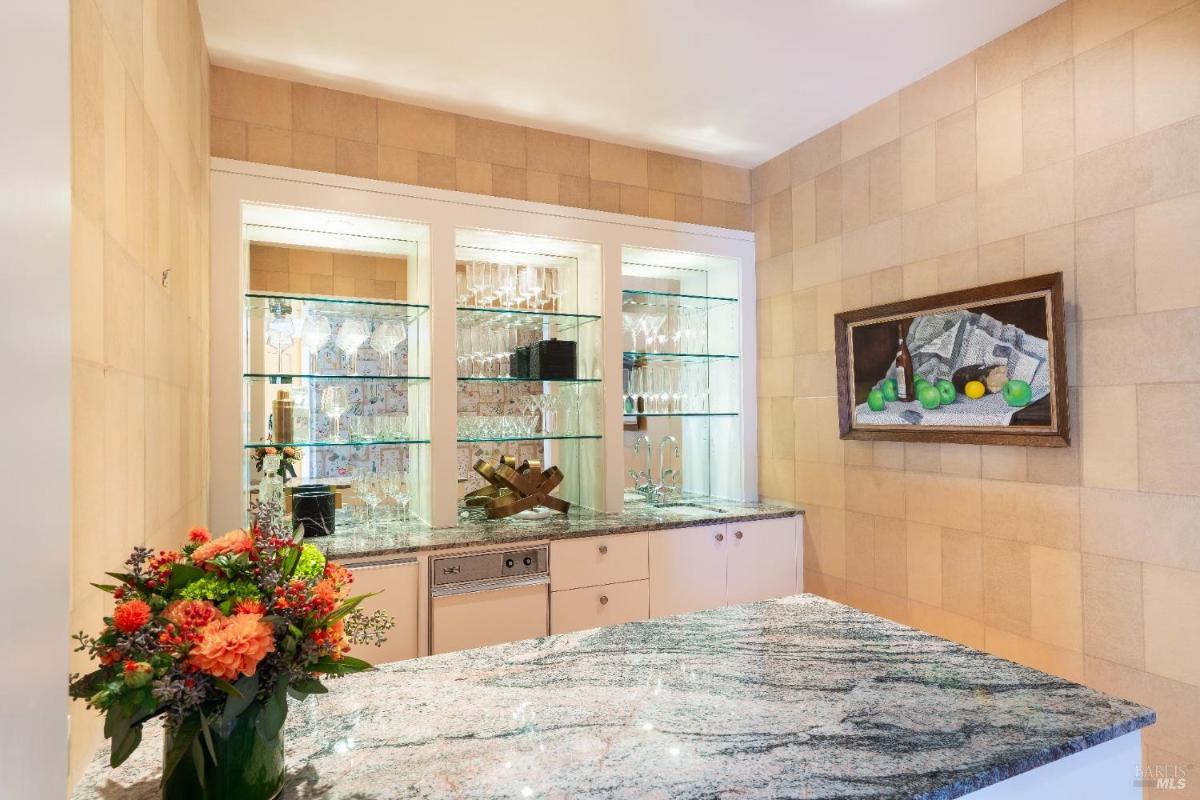 Bar with granite countertop with glass shelves displaying glassware and a framed still-life painting.