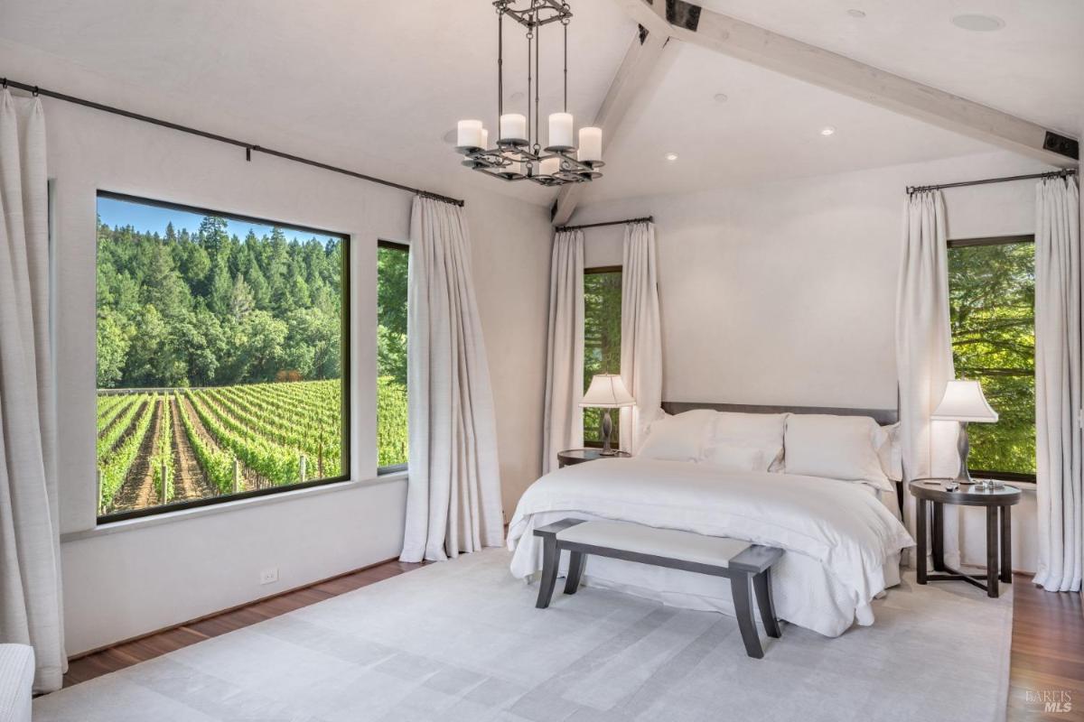 A bedroom with a bed and two bedside tables, featuring large windows that overlook a vineyard and forest.