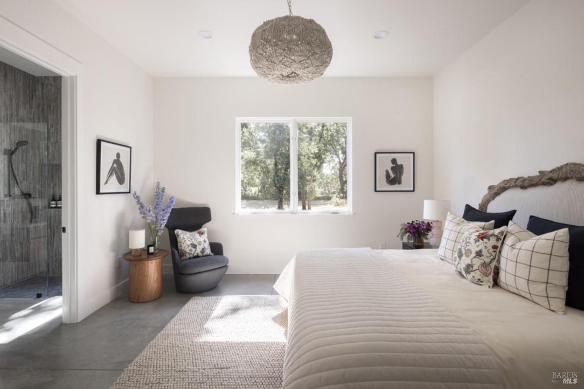 A bedroom with a large bed, a sitting area with a gray armchair, and an en suite bathroom visible through an open doorway.