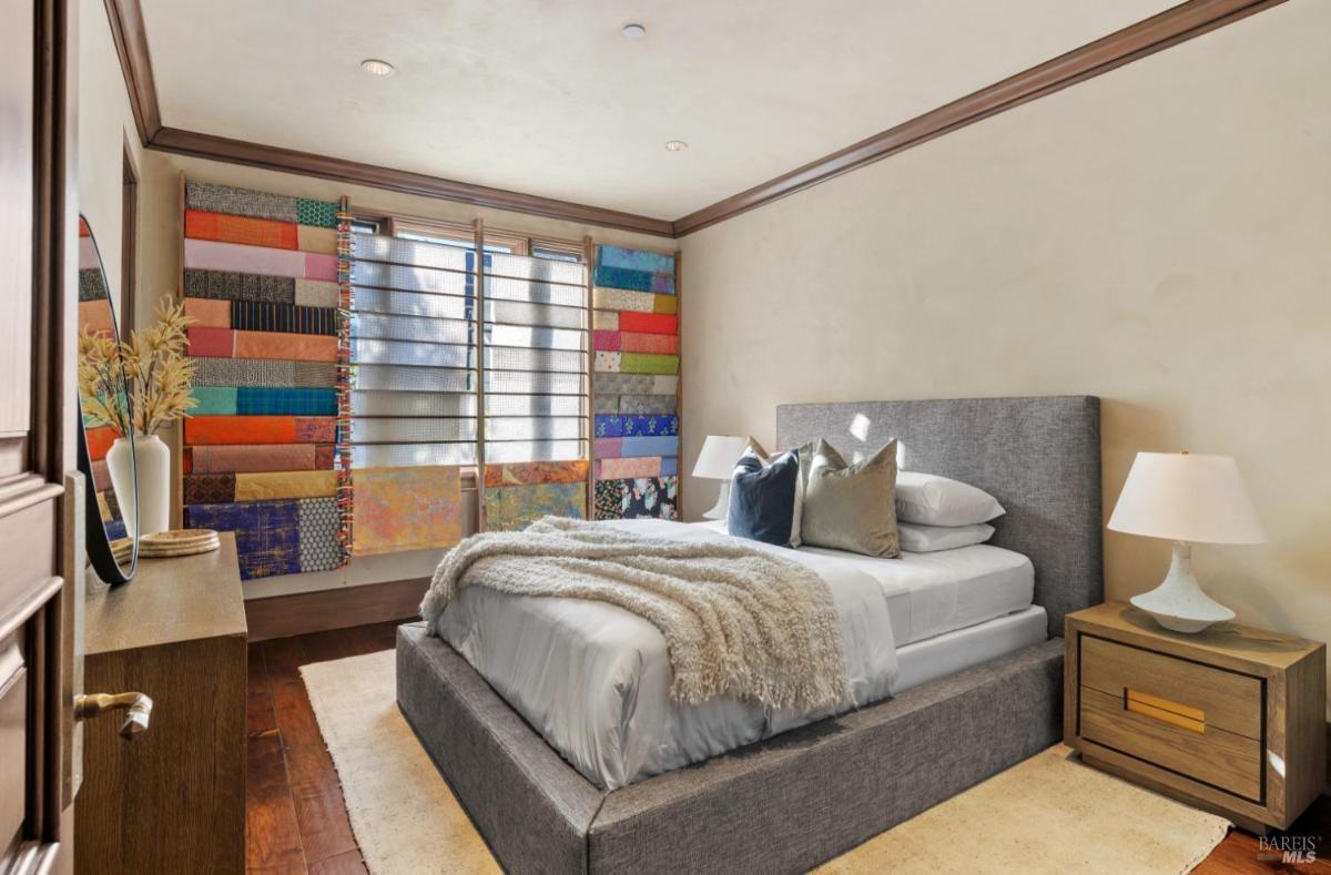 Bedroom featuring a gray upholstered bed with light blue bedding. The room has colorful patterned curtains and two bedside lamps. 