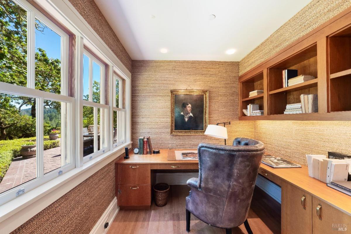 Office desk with bookshelves, a framed portrait, and windows overlooking greenery.