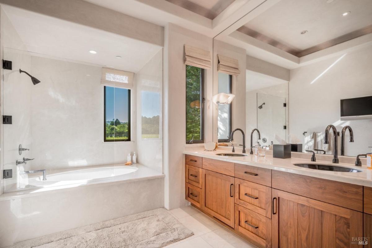 A bathroom with a bathtub, shower, two sinks, and a large mirror, featuring windows that provide a view of the outdoors.