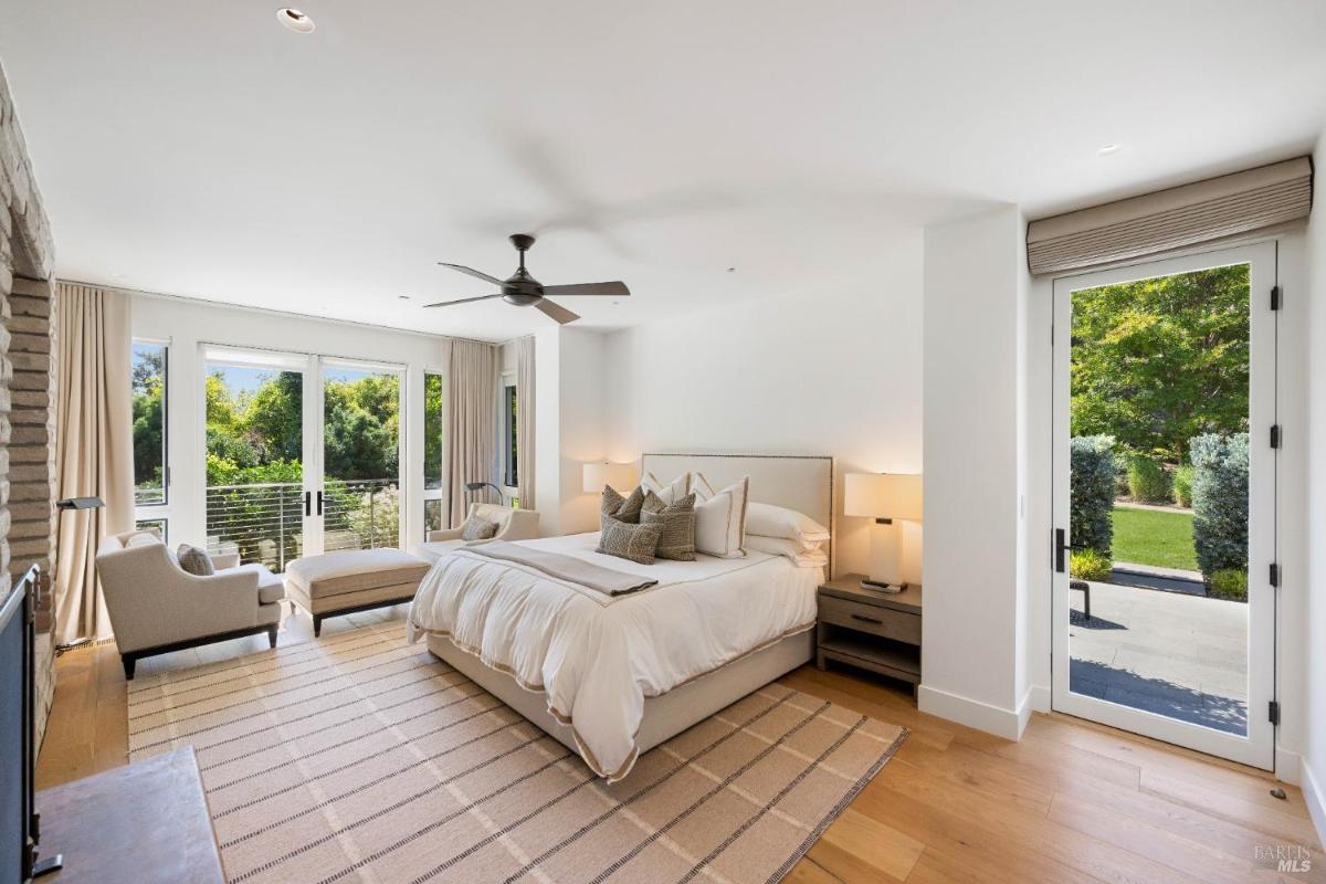 A bedroom with a bed, bedside tables, a ceiling fan, chairs, and glass doors opening to a garden.