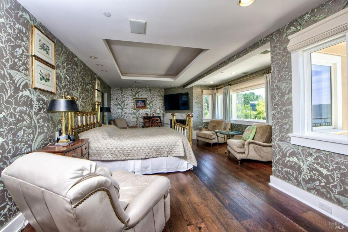 Bedroom with patterned wallpaper, a brass bed frame, and a sitting area by large windows overlooking greenery.