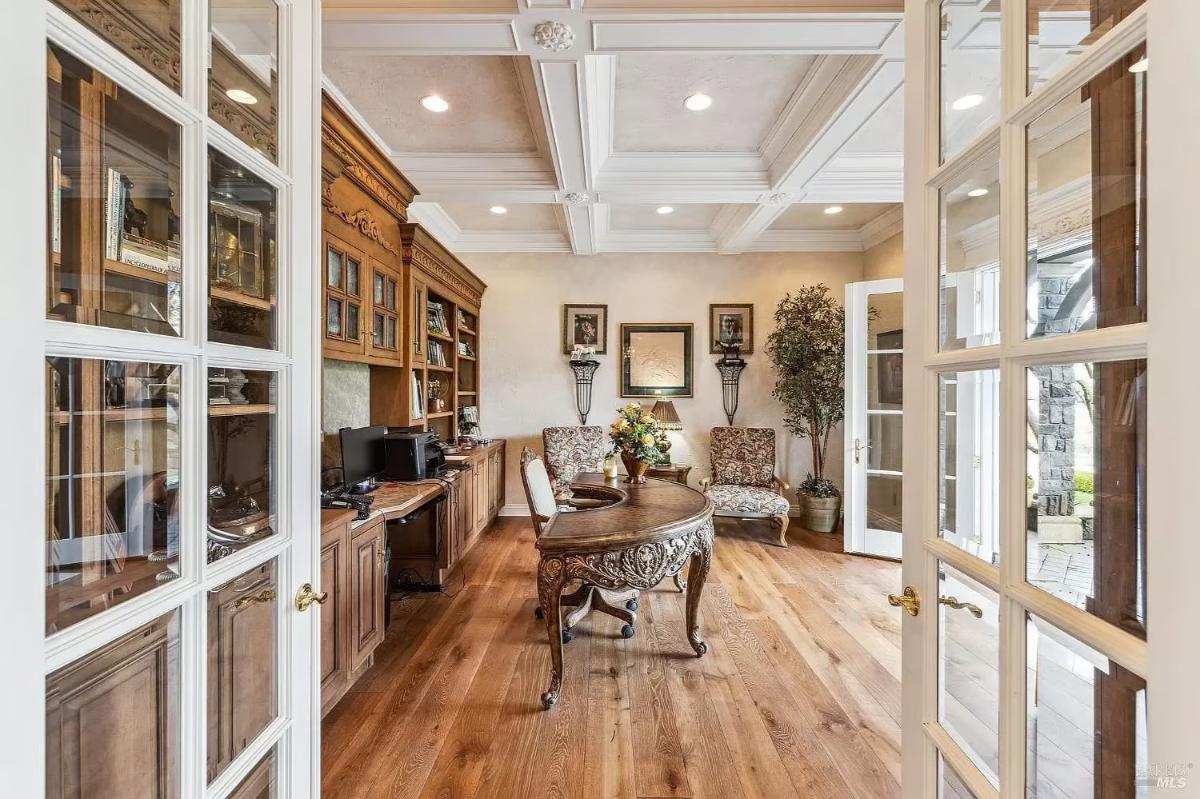 Home office with a large wooden desk, built-in bookshelves, and a coffered ceiling, accessible through French doors.