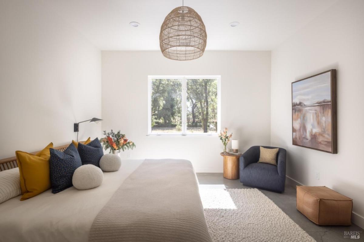 A bedroom with a large bed, colorful pillows, a seating area with a dark chair, and a window overlooking greenery.