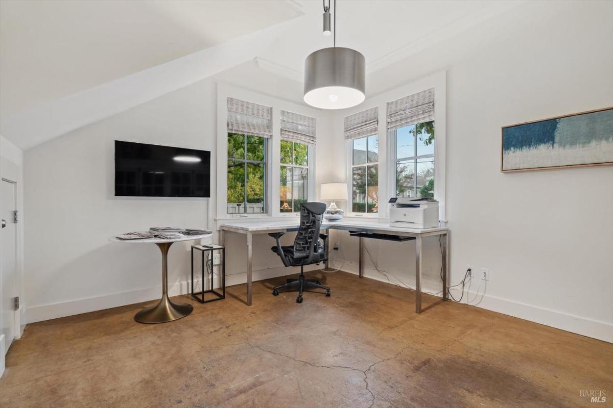 A home office with desk with a chair and a television on the wall near large windows.