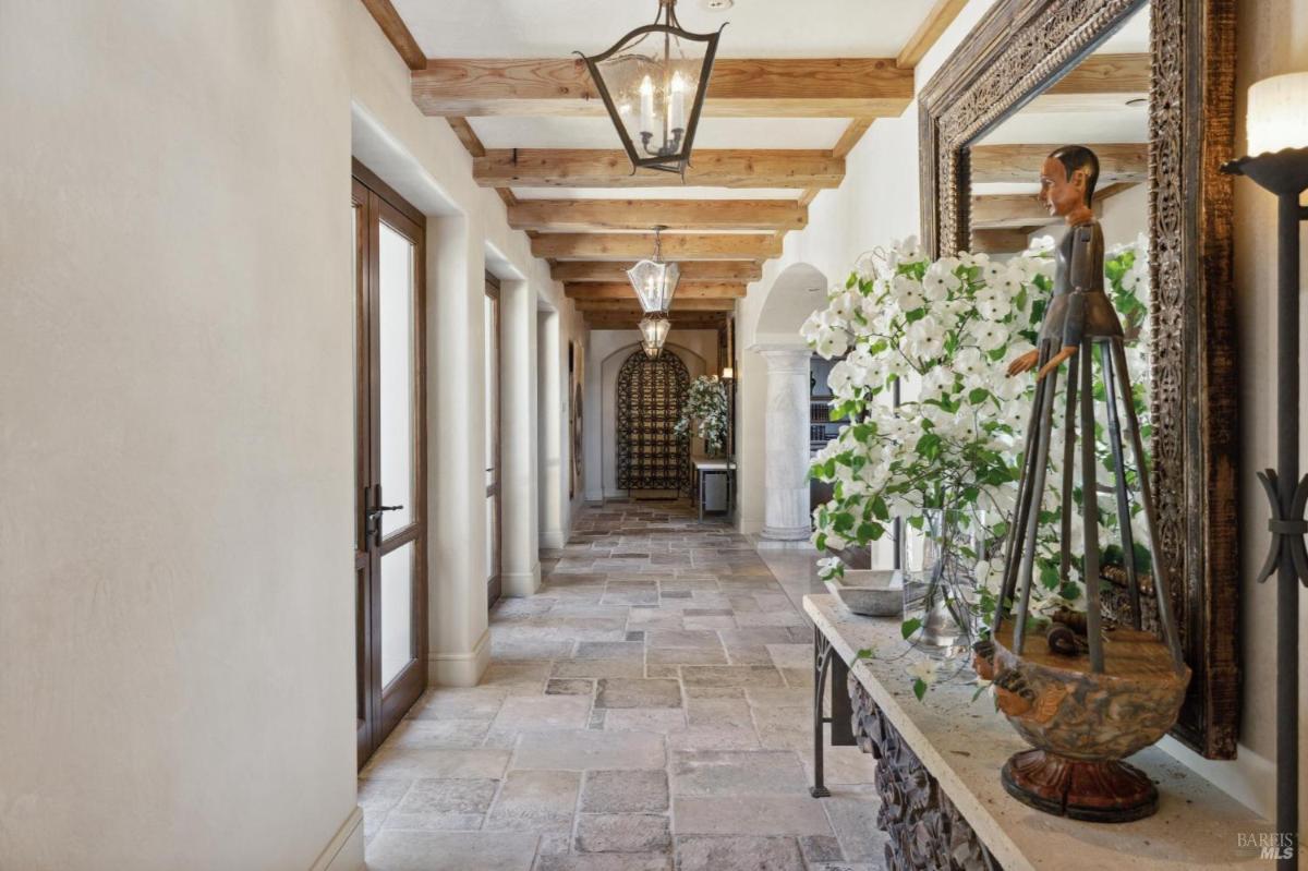 Hallway with wooden beams on the ceiling, the stone flooring, and decorative features like the statue and floral arrangements create a warm and inviting atmosphere.