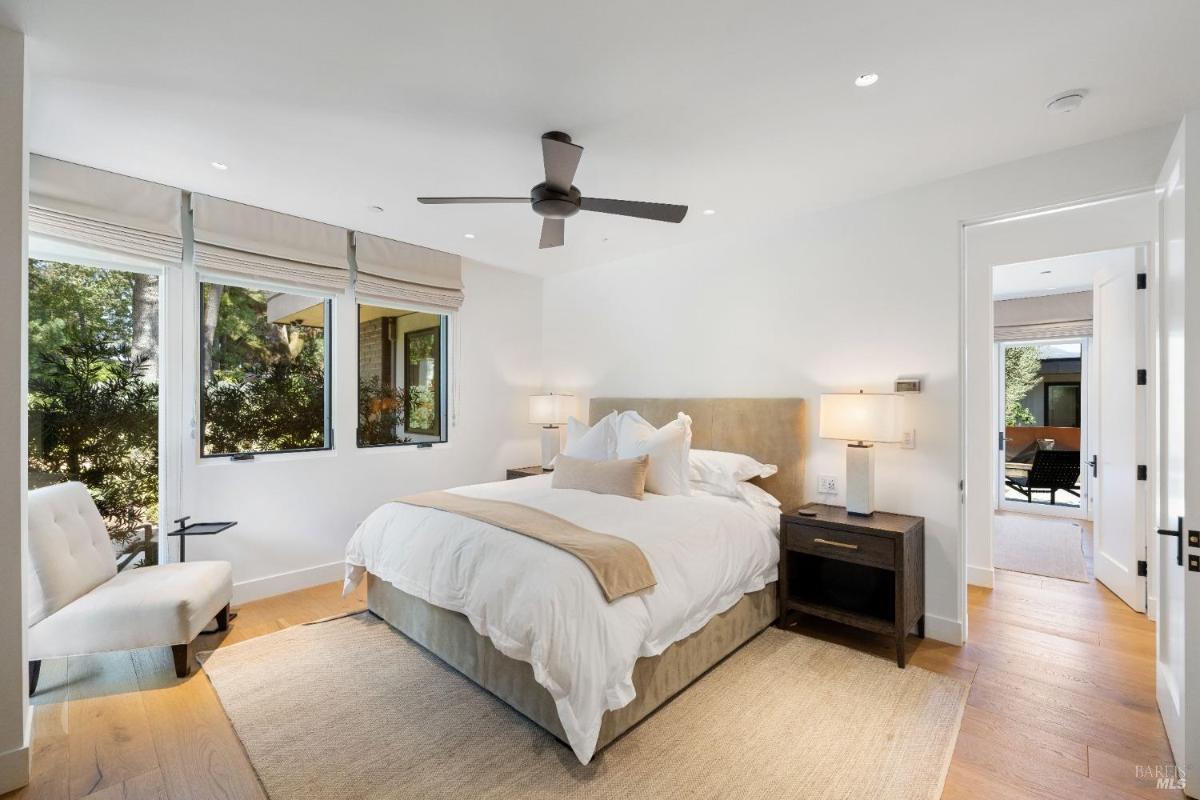 A bedroom with a bed, bedside tables, a chair, and windows overlooking greenery.