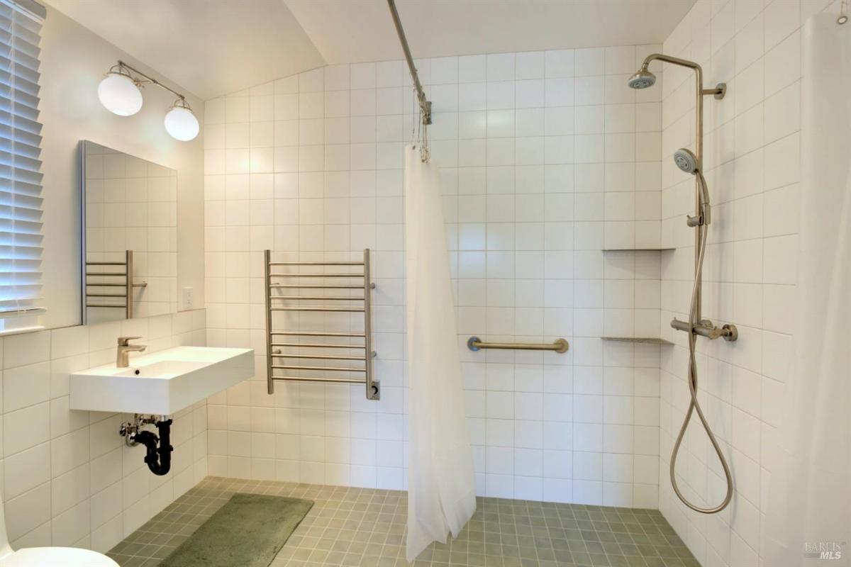 A bathroom with white tiled walls, a walk-in shower with handrails, a sink, and a heated towel rack.