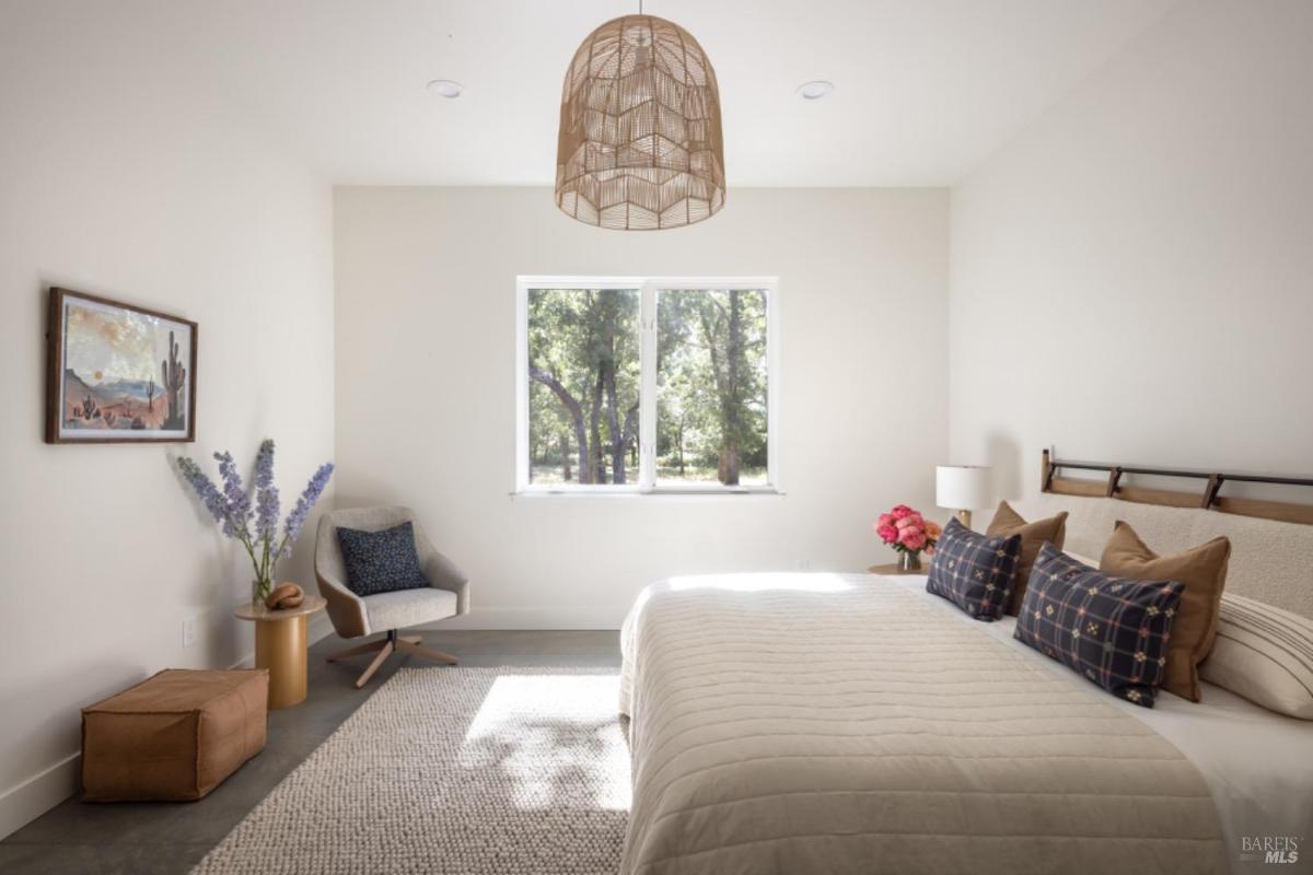 A bedroom with a large bed, seating area, decorative pillows, and a window with a view of the trees outside.