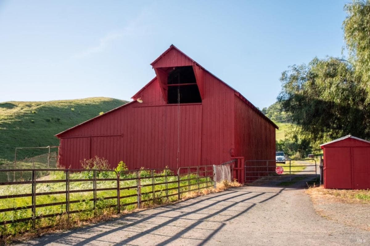 Red barn with a fenced driveway and green hills in the background.