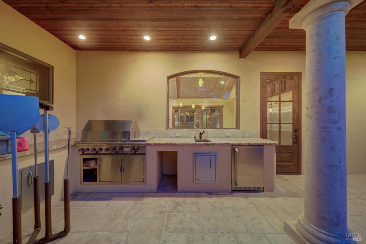 An outdoor kitchen featuring a grill, counter space, a sink, and a refrigerator, with wooden ceiling beams and stone columns.