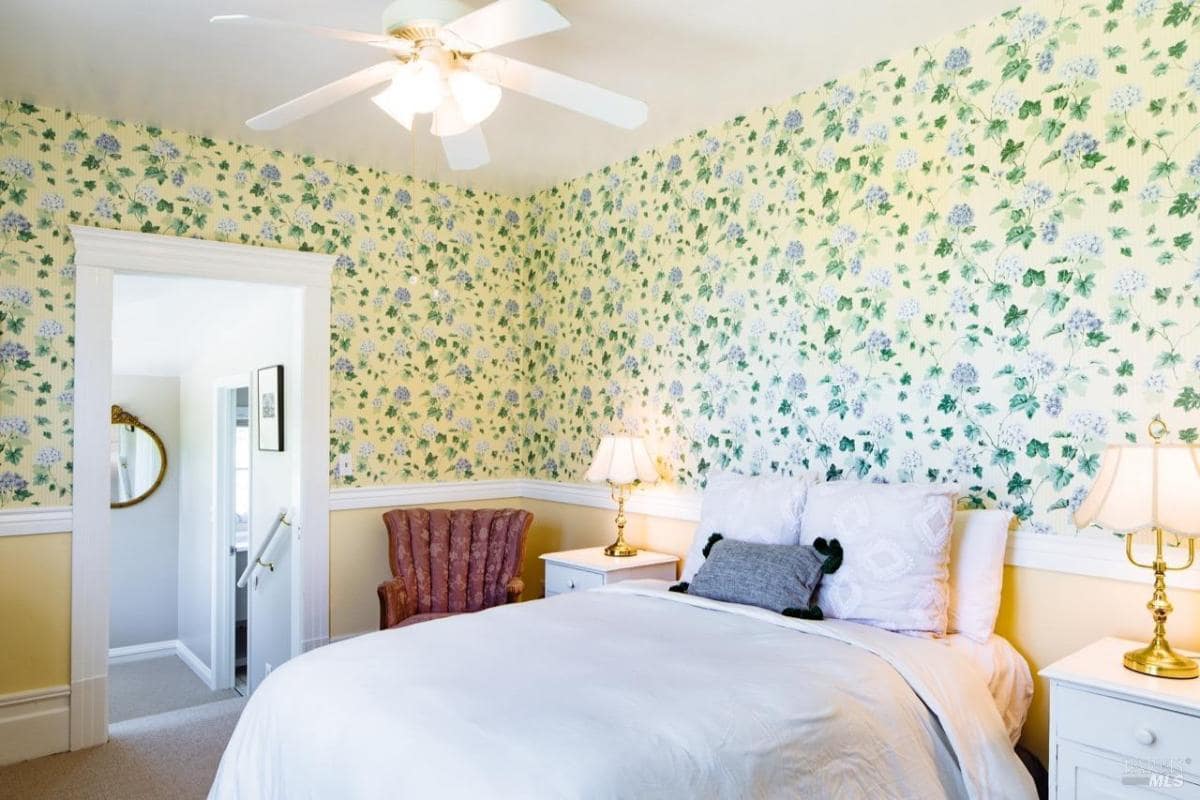 A bedroom with floral wallpaper, a double bed, and a chair near the window.