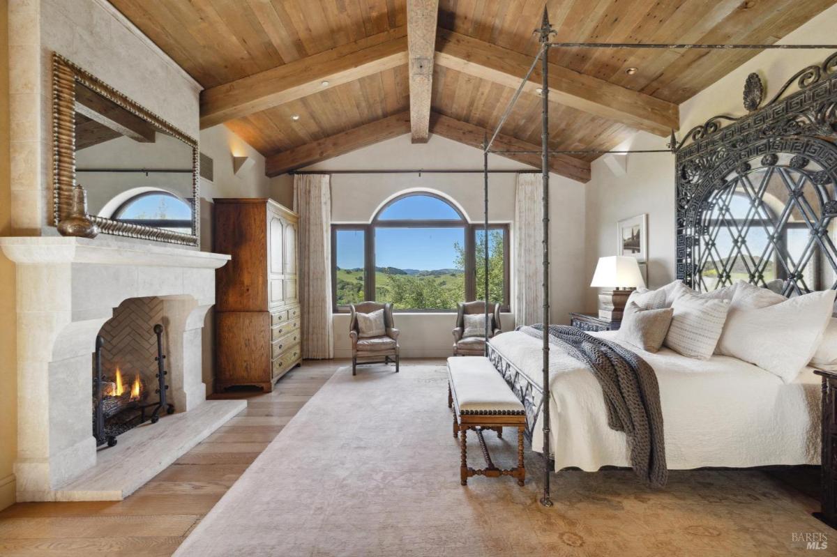 A bedroom with a four-poster bed, a fireplace, and a large window showing a view of the outdoors.