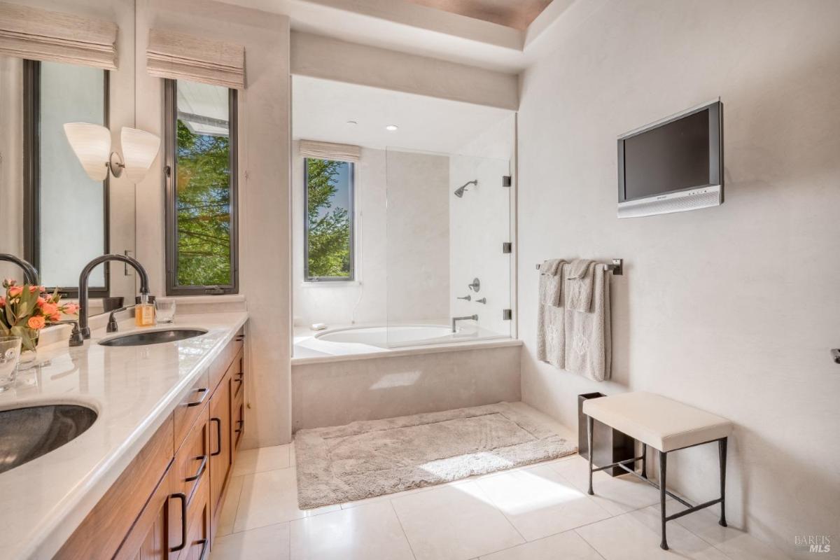 Bathroom with a glass shower, a bathtub, dual sinks, and a wall-mounted TV.