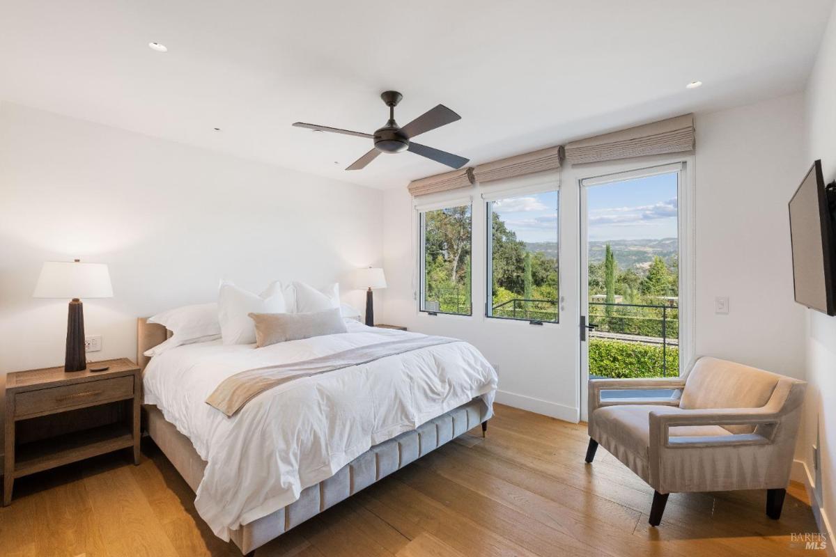 A bedroom with a bed, bedside tables, a ceiling fan, and windows with a view of a landscaped garden.