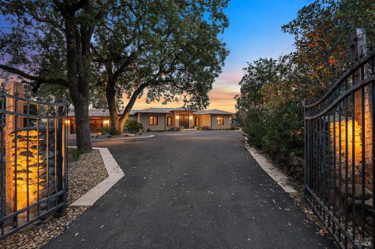 A gated driveway entrance with a lit stone pillar and trees.