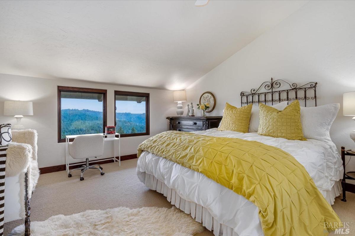 A bedroom with a bed featuring yellow bedding, a desk with a chair near windows, and carpet flooring.