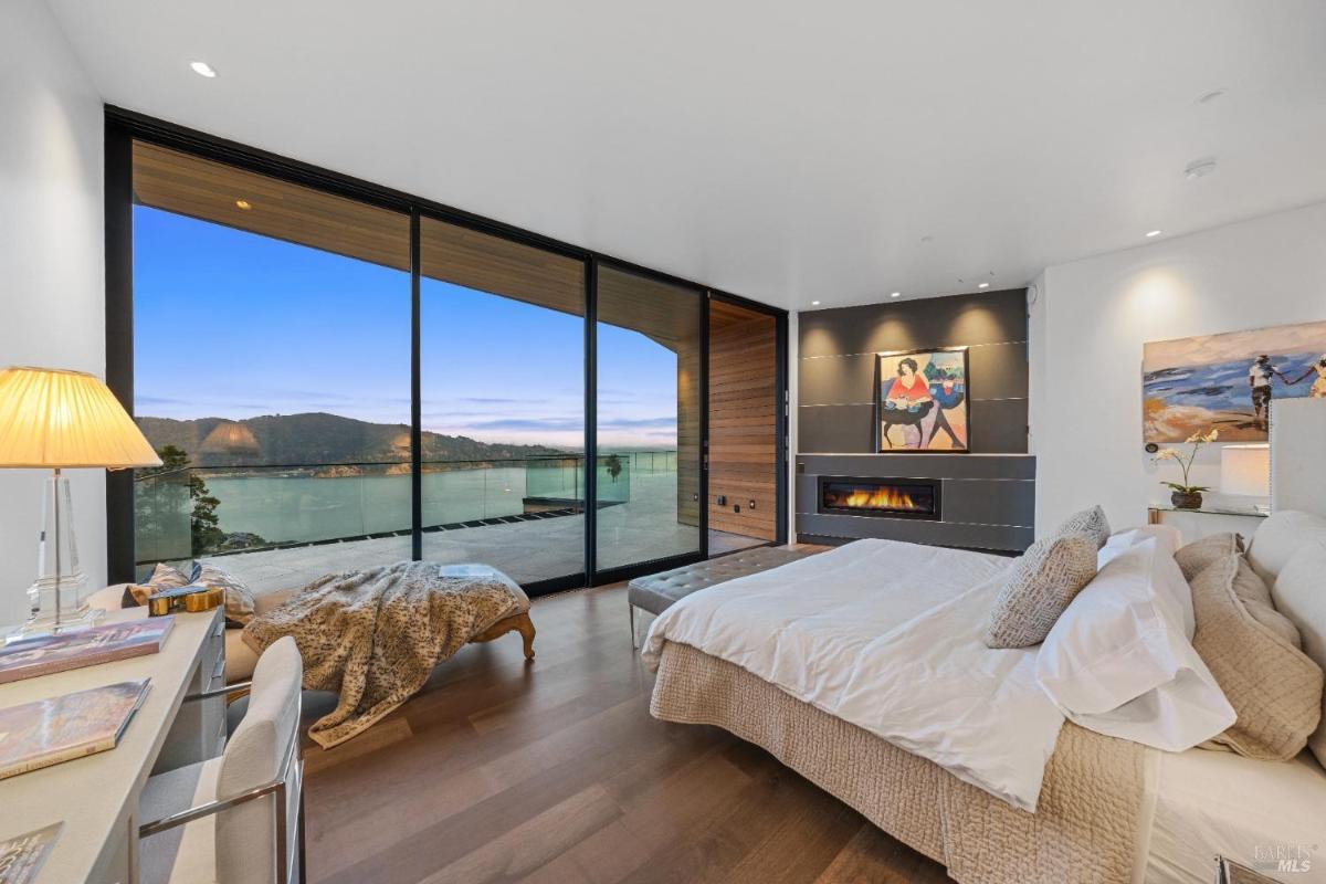 A bedroom with a large glass wall overlooking a bay, a fireplace, and a large bed.