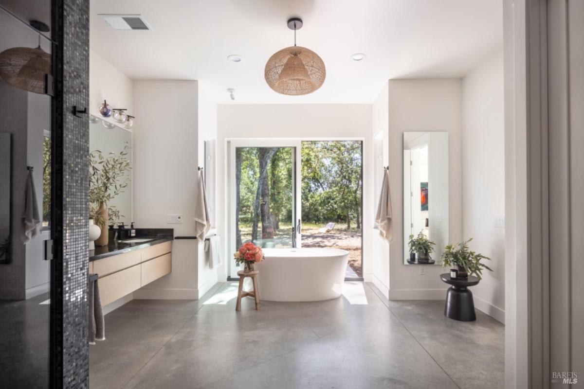 A spa-inspired bathroom with a freestanding bathtub, large windows overlooking trees, dual vanities, and modern fixtures.