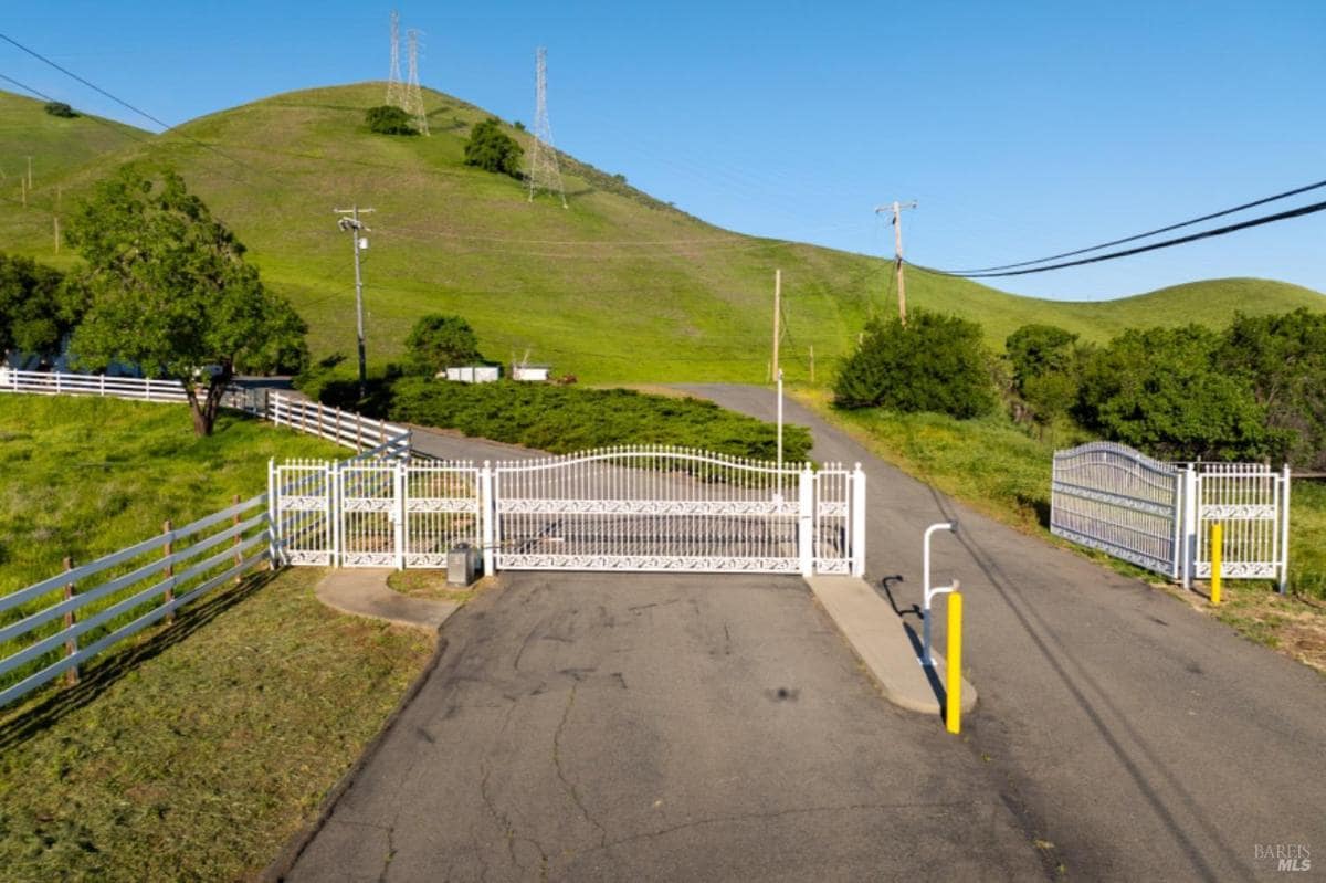 White gated entrance with paved road leading to green hills.