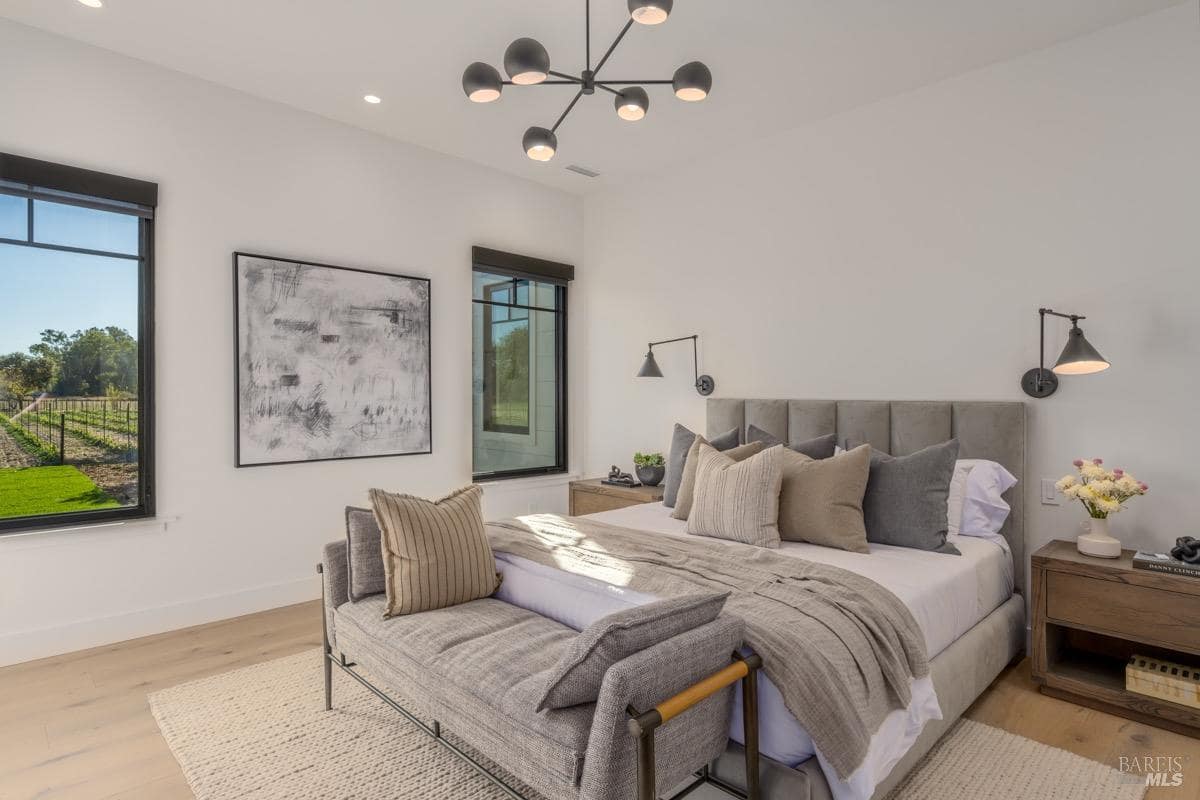 A bedroom with a large upholstered bed, bench seating, wall-mounted lights, and windows overlooking the outdoors.