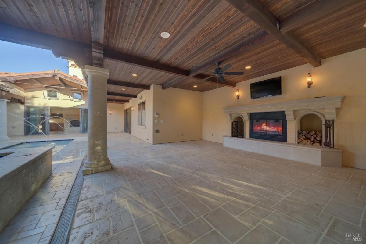 An outdoor living area with a stone fireplace, wooden ceiling beams, and a view of a pool. 