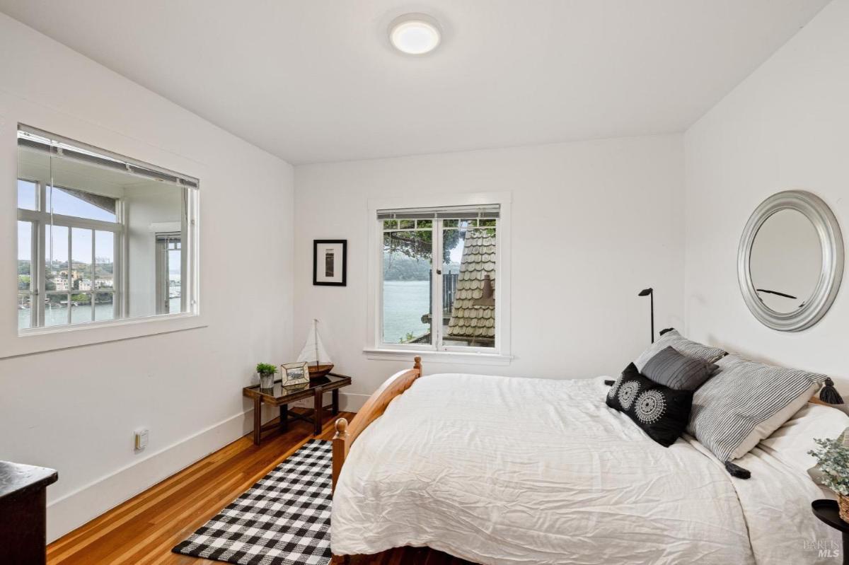 A small bedroom with white walls, a single bed, and a view of a water body through the window.