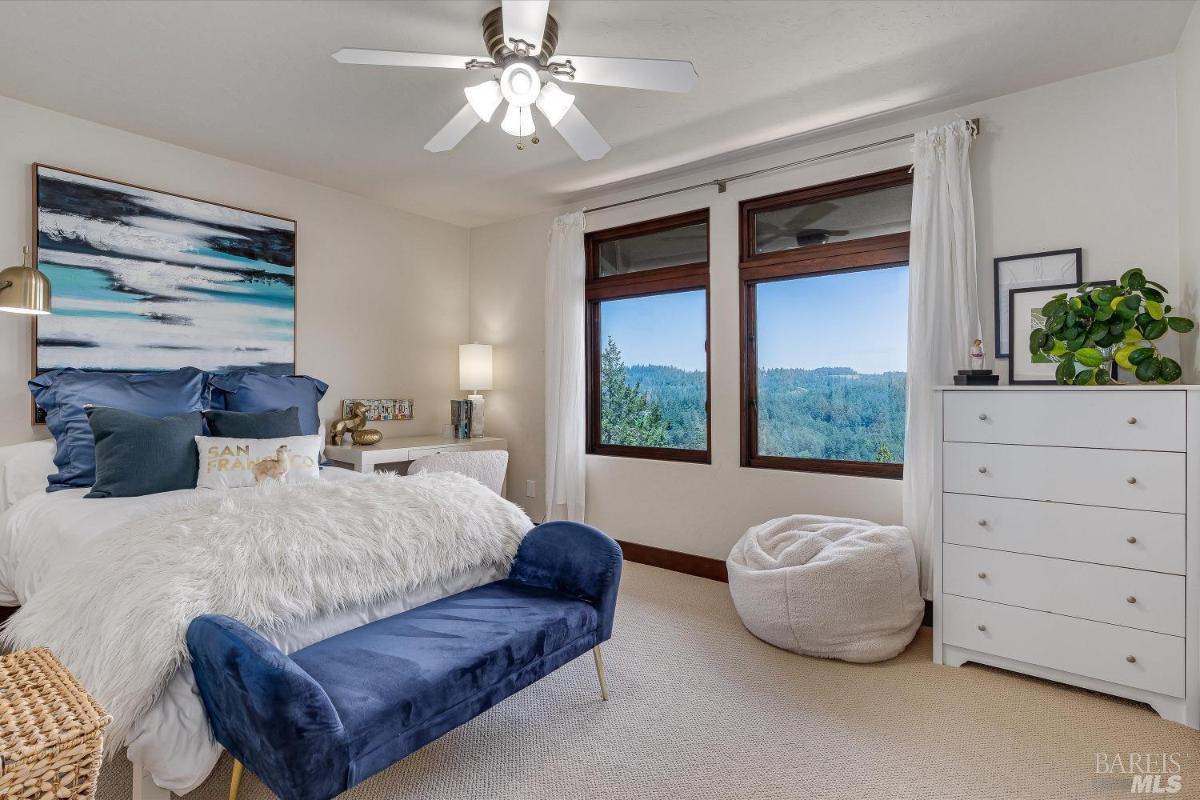 A bedroom with a bed, blue bench, dresser, and windows overlooking a view.