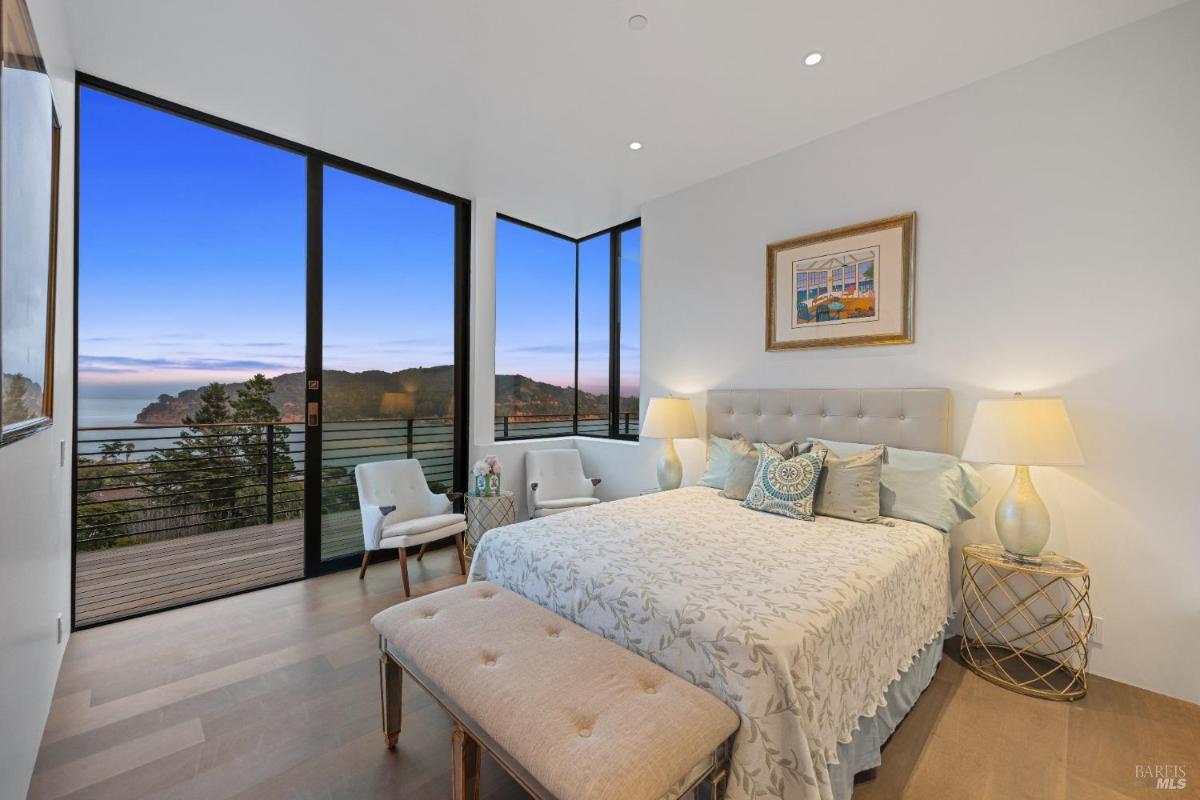 A bedroom with large windows overlooking a bay, a large bed, and a small seating area.