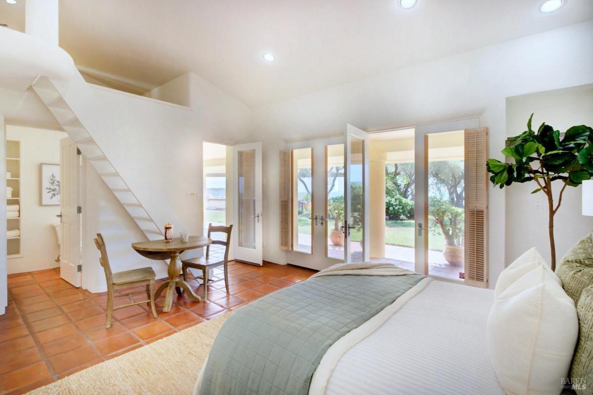 A bedroom with loft stairs, a small dining table, and doors opening to a garden.