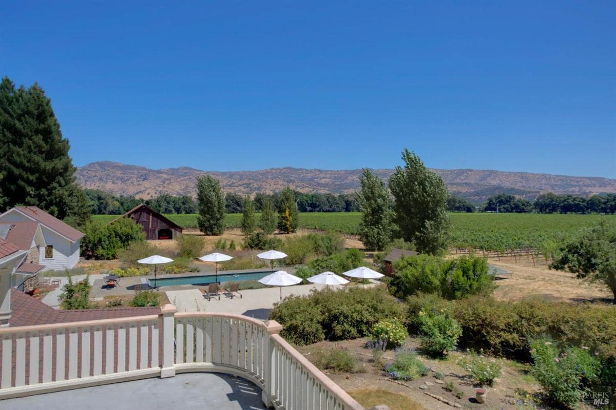 A balcony overlooking a yard with a pool, seating area, and a barn in the background with a vineyard view