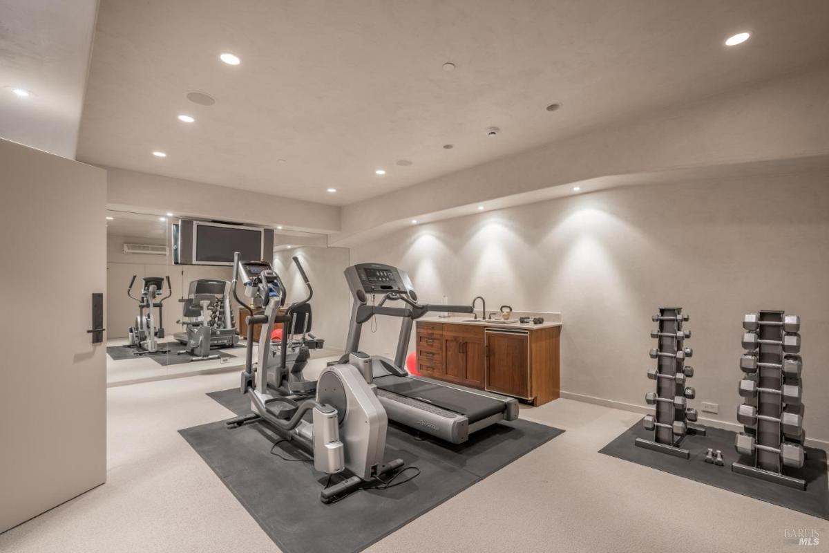 A gym interior featuring a treadmill, an elliptical machine, a weight rack with dumbbells, and a wall-mounted TV.