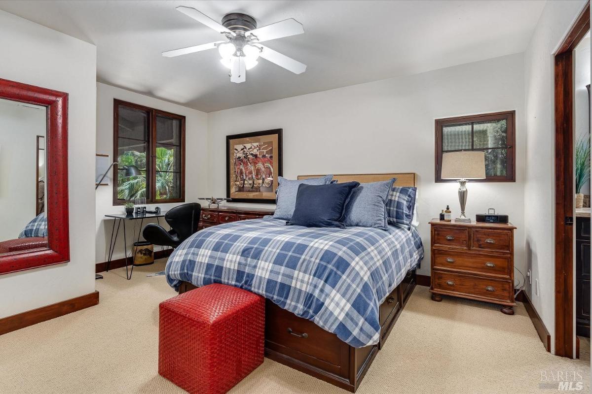 A bedroom with plaid bedding, a desk, a red ottoman, and a ceiling fan.