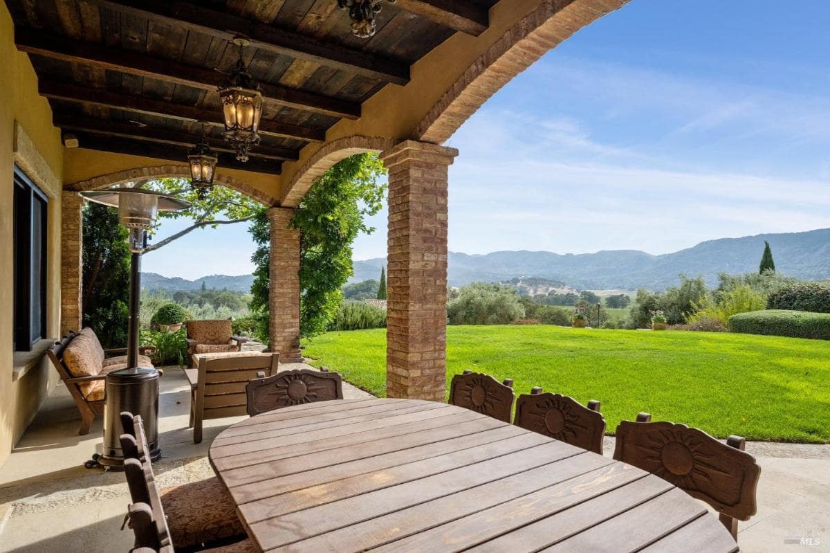 A covered outdoor patio with wood beams, a dining table, seating, and a view of a green lawn and hills.