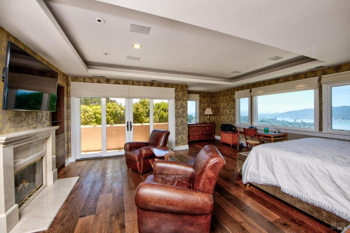 A bedroom with large windows, a fireplace, seating, and balcony access.