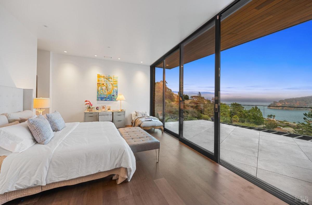 A bedroom with a bed, a bench, and a large glass door opening to a deck with a bay view.