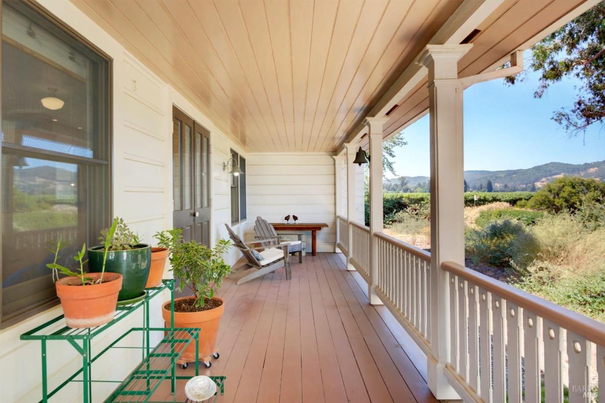 A covered wooden porch with railings, potted plants, and chairs, with a view of greenery and hills in the distance.