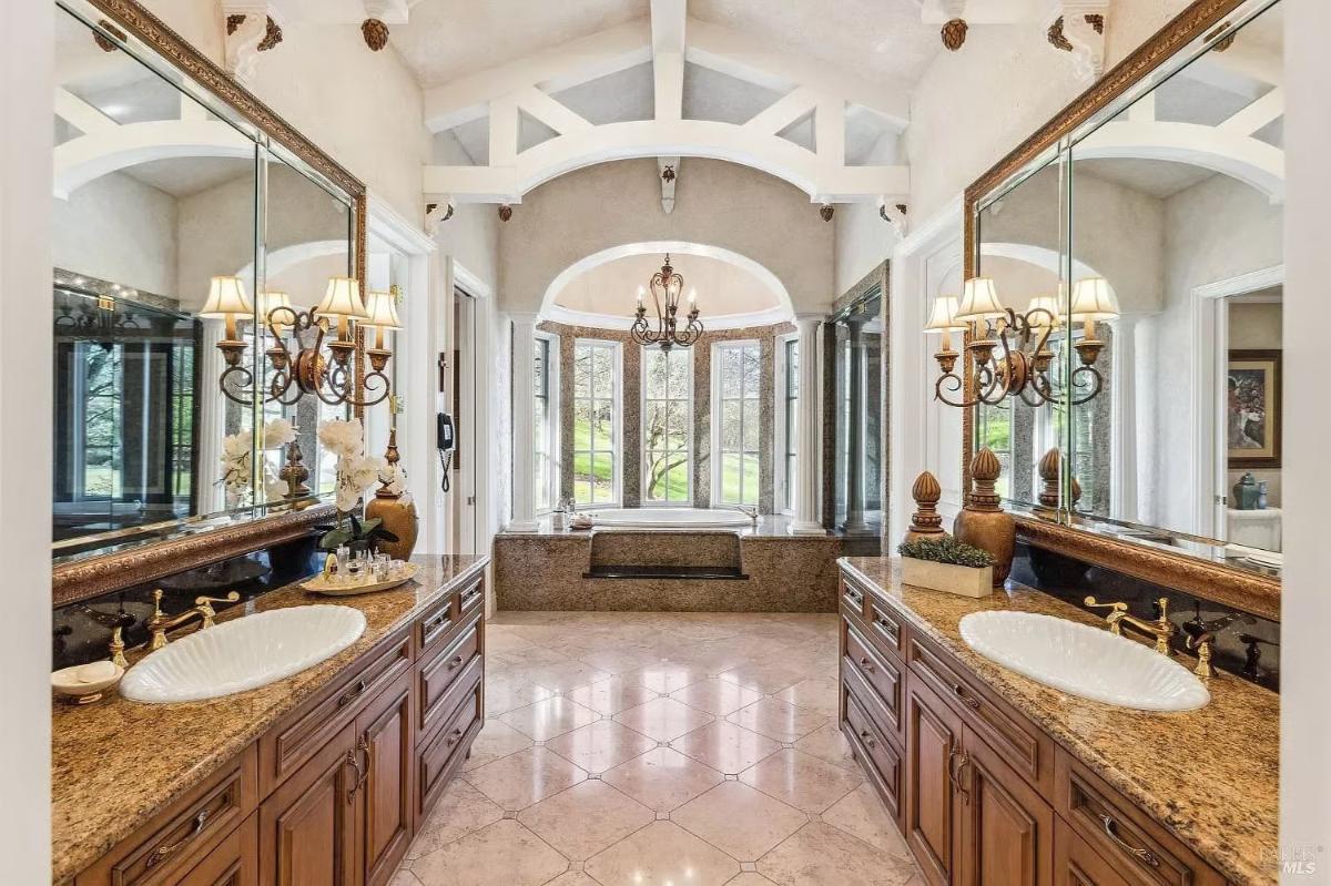 Bathroom with double vanities, granite countertops, decorative mirrors, and a central soaking tub surrounded by arched windows.