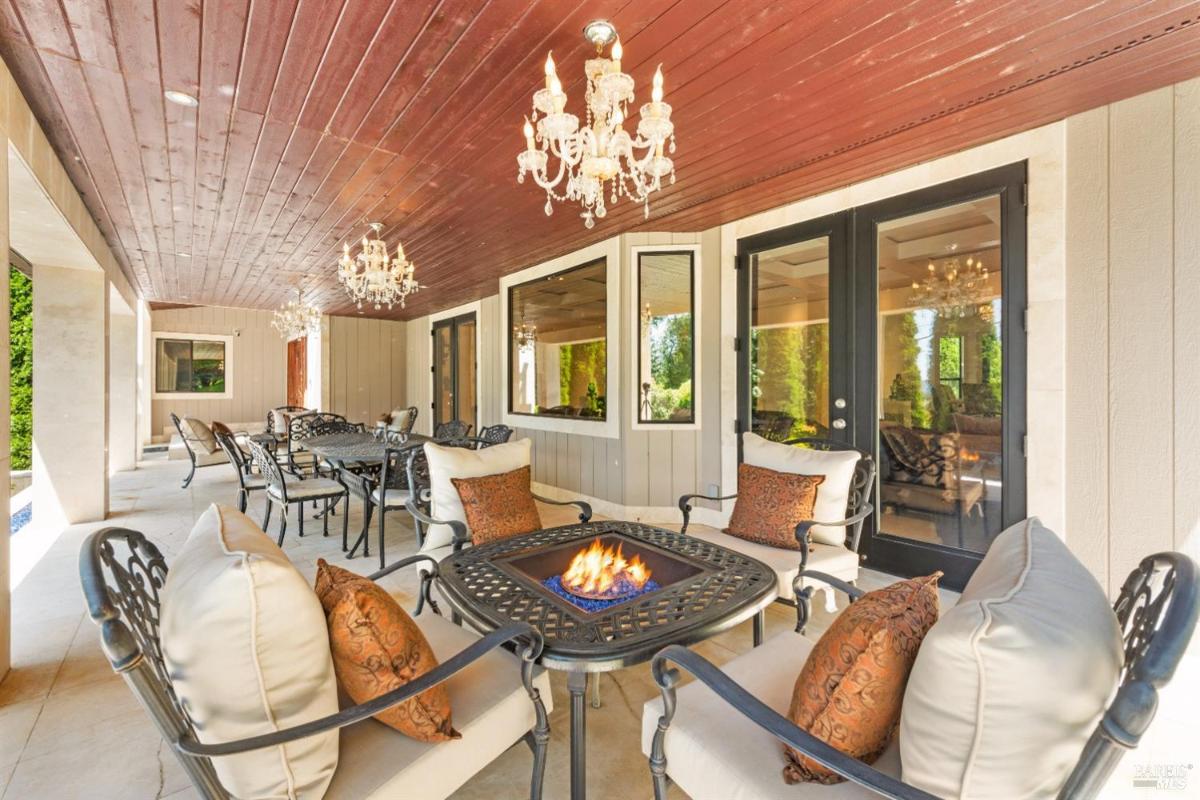 A covered patio with seating, a fire pit, and chandeliers. Large windows provide views of the outdoors.