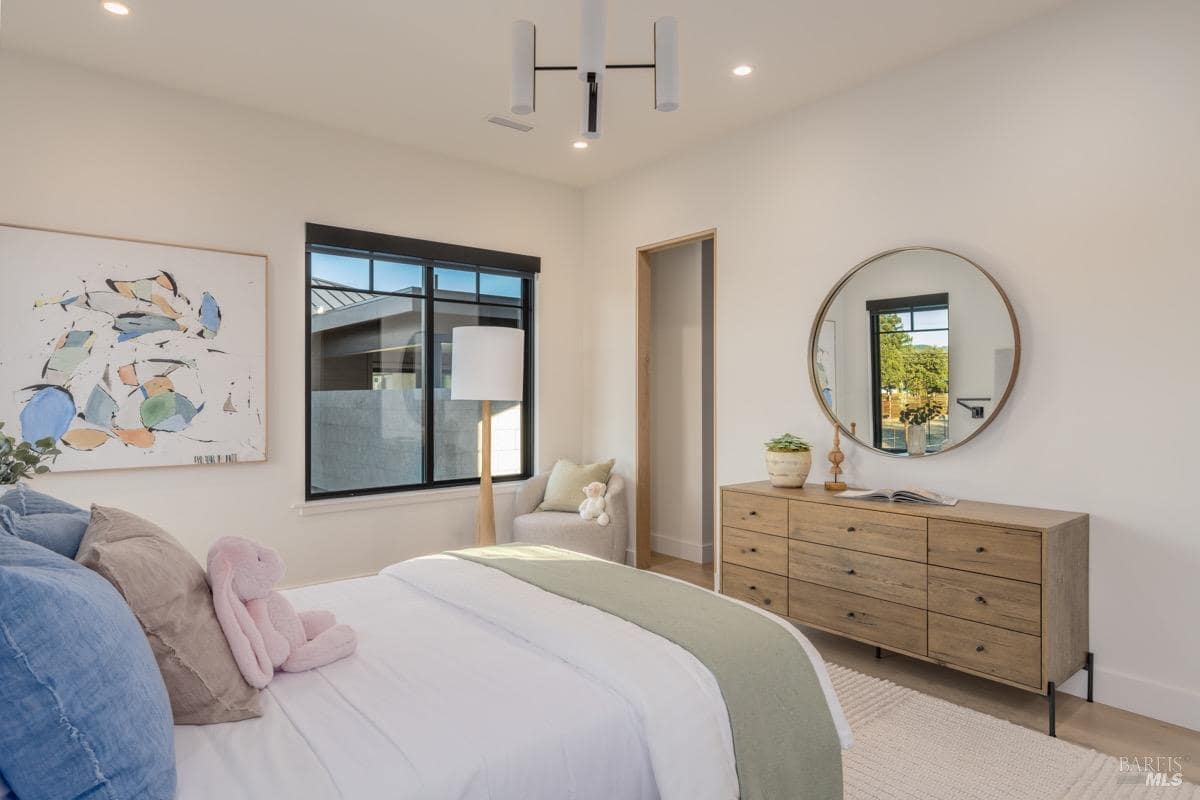 A bedroom with a large bed, wooden dresser, round mirror, abstract wall art, and a window with natural light.