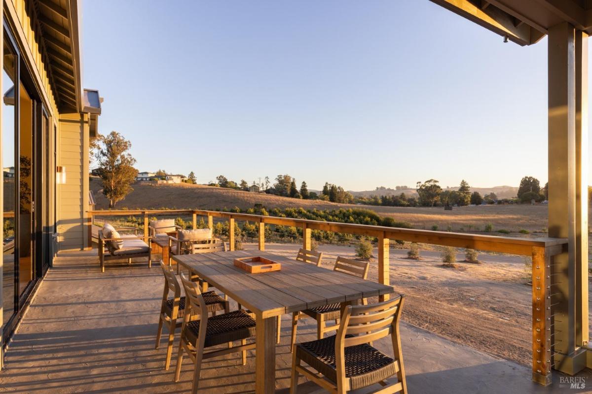 A wooden deck with a dining table and chairs overlooks a grassy landscape at sunset.