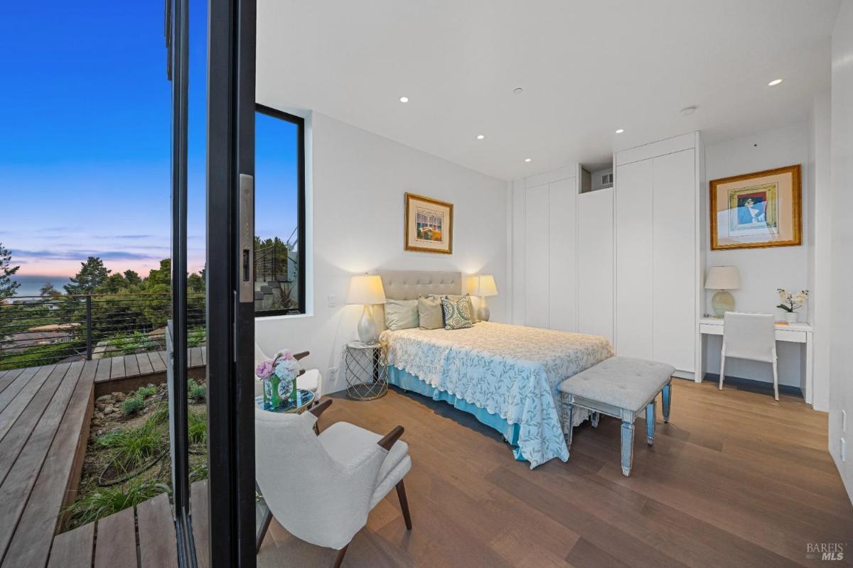 A bedroom with a bed, white built-in wardrobes, a small desk, and a sliding door overlooking a deck and bay.