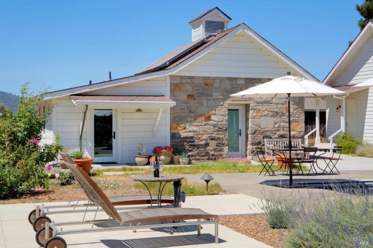 A stone and white-paneled outbuilding with outdoor lounge chairs, a table, and umbrellas in a garden setting.