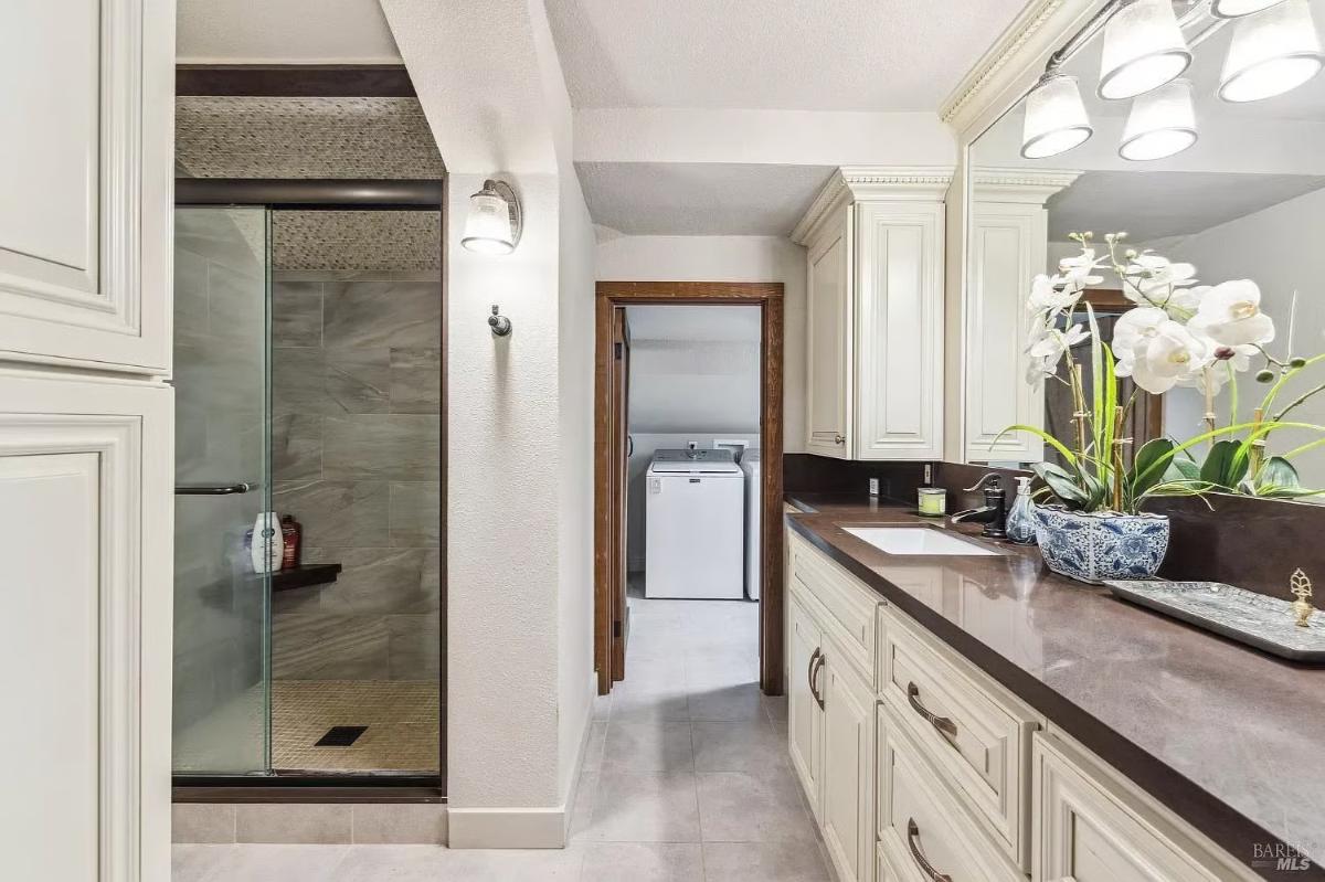 Bathroom with a glass-enclosed shower, laundry area in the background, and a single vanity with cabinets.