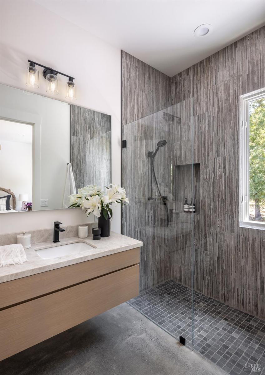 A bathroom with a glass-enclosed shower featuring textured gray tile, a floating vanity with a light countertop, and a large mirror.